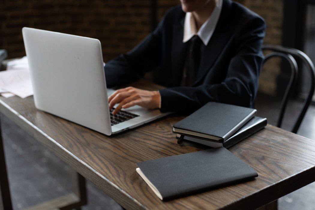 A person working at a laptop on a wooden desk beside a notebook and a mug